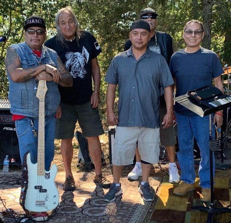 Five men, all Native American, stand wearing contemporary clothing in a group photo. One holds a bass guitar.