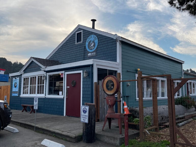 A wood building painted dark blue with a red door and four windows facing to the left of the camera lens. A wooden dock surrounds the building. A sign on the building reads "Slack Tide Cafe."