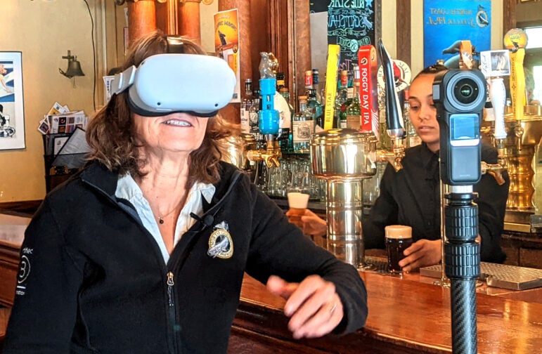 A woman sits at a bar, facing the camera, wearing a black fleece jacket and wearing a white virtual reality headset.