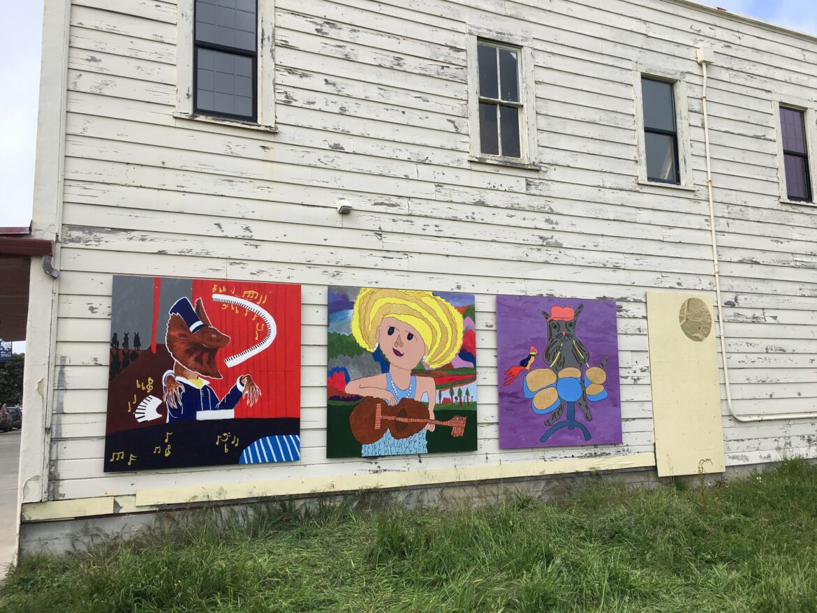 Three square shaped murals hang on a white wooden wall of a building. The murals depict a lizard playing piano, a woman with blonde hair playing guitar, and a cat playing drums.