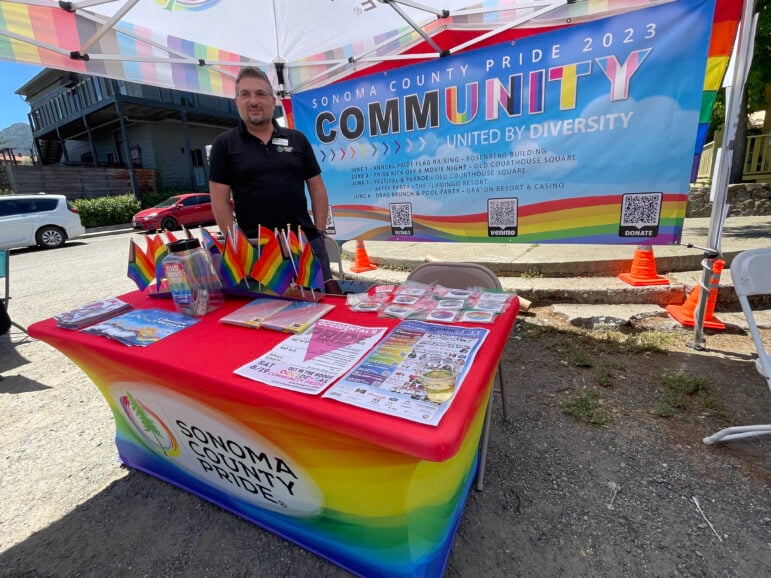 A man stands behind a table decorated with LGBT pride rainbow colors.