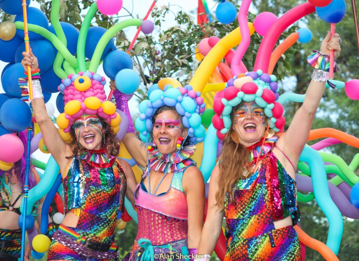 Three cheering women stand together wearing rainbow colored clothing and hats made of colorful balloons.