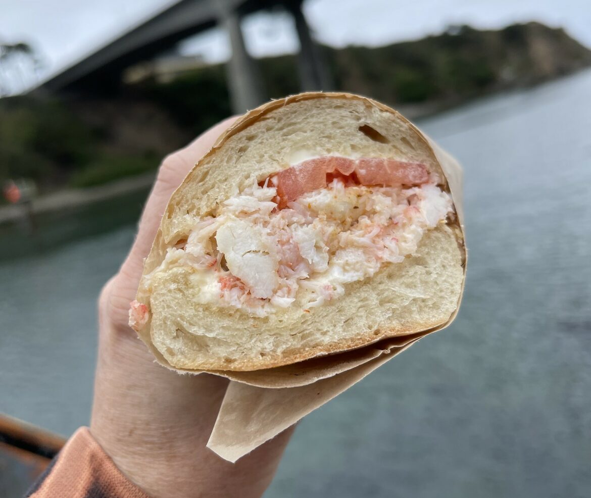 A hand holds a Dungeness crab sandwich. The sandwich comprises two pieces of bread. Between the bread is fresh crab and a single tomato.