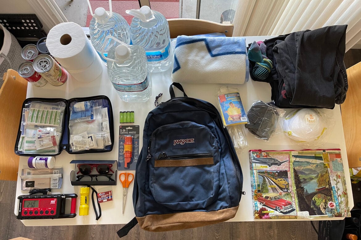 Emergency supplies for a go bag, including a medical supply kit, important papers, poncho, blankets, clothing, a flashlight, eyeglasses, face masks, emergency radio and more. Also pictured is bottled water, canned goods and toilet paper for a go box.