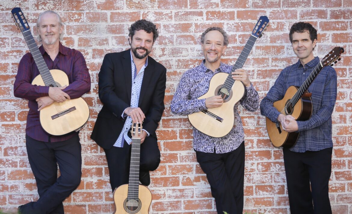 A photograph of four men standing against a brick wall. All are smiling and holding acoustic guitars.