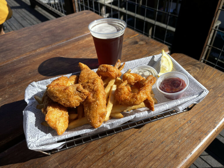 A plate with fried rock cod fish, fried shrimp, french fries, a container of ketchup and tartar sauce and a beer on the side.