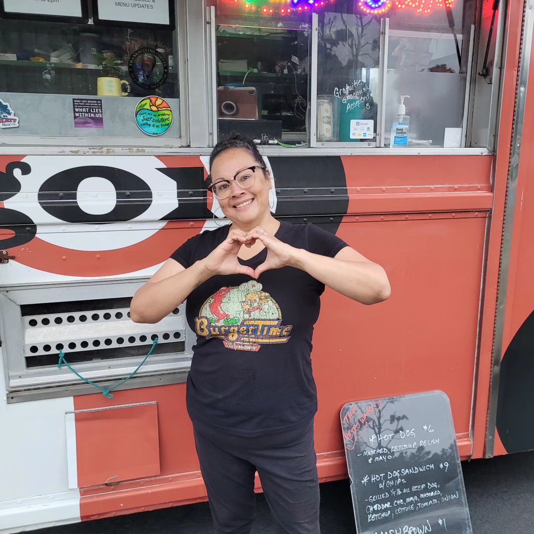 A woman wearing a black t-shirt and black pants and eyeglases stands with her hands in a heart symbol in front of a food truck.